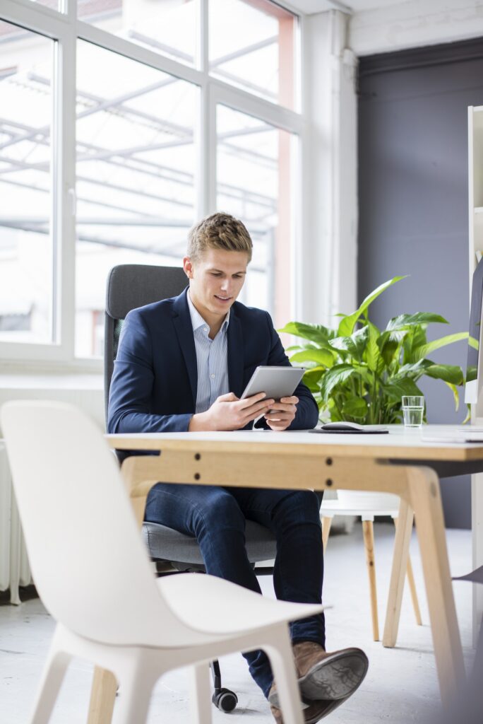 Confident young businessman sitting at desk in office using tablet