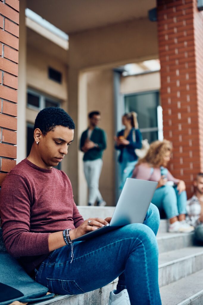 Young black student using laptop while relaxing on the steps of university building.